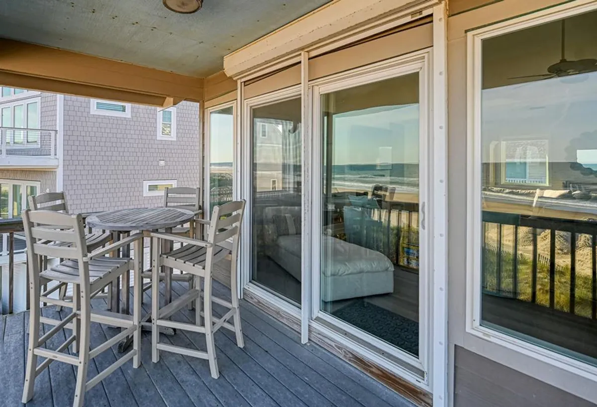 A serene exterior scene featuring an outdoor dining set facing the vast ocean. The area is framed by large windows, integrating indoor comfort with outdoor relaxation. The gentle morning light suggests a perfect spot for sunrise breakfasts or evening gatherings with the sound of ocean waves close by.