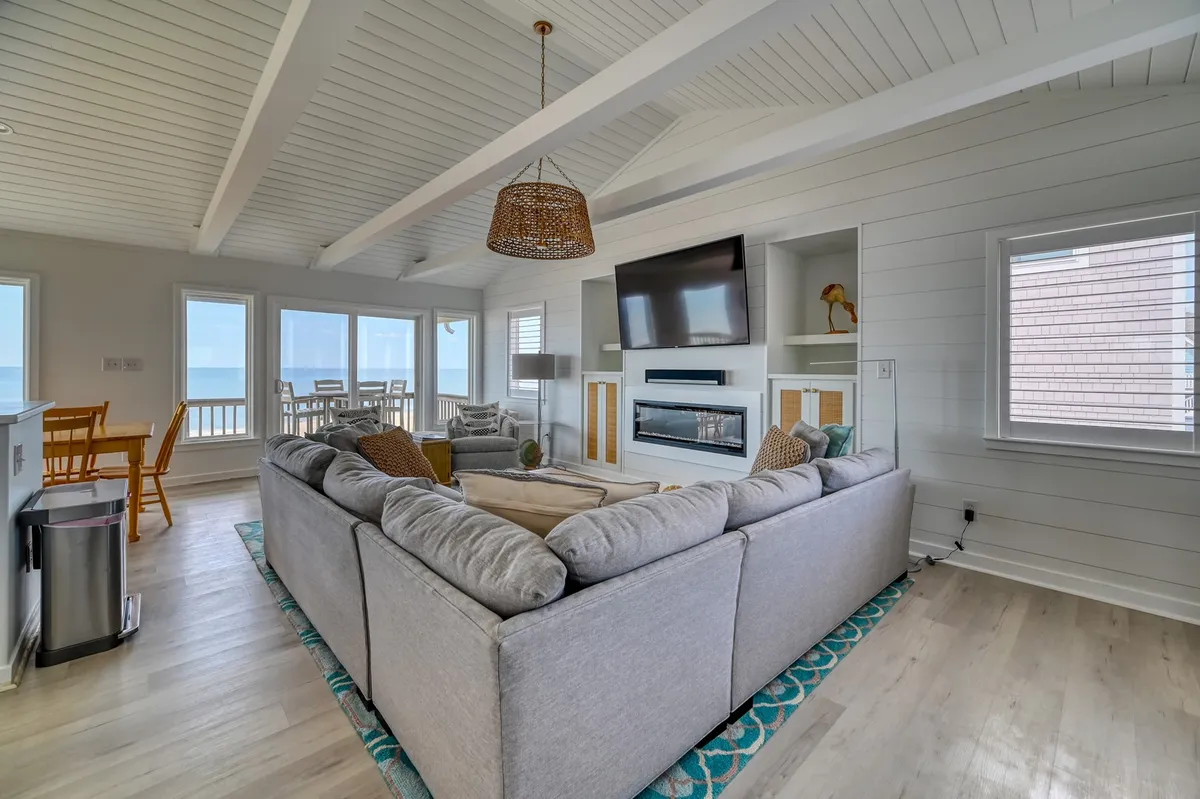 This image features a spacious Great Room with a large sectional sofa, a cozy fireplace, expansive windows showcasing the ocean, and a TV mounted on the wall. The room exudes a sense of comfort and openness, bathed in natural light with a clear view of the Atlantic.