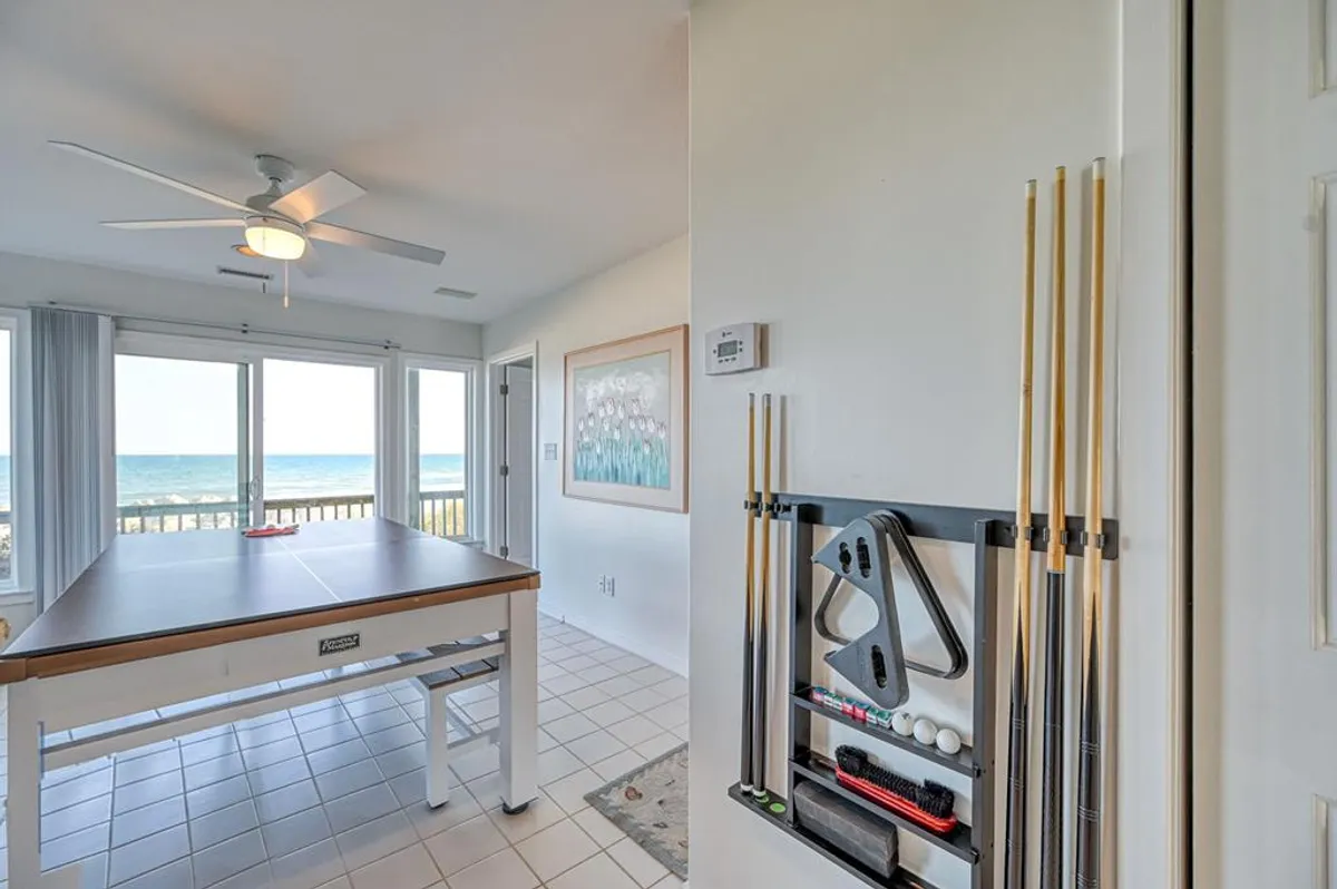 An interior game room featuring a pool table positioned in front of large windows with ocean views. The space is bright and inviting, with artwork and comfortable surroundings that suggest playful fun and memorable moments for guests of all ages.