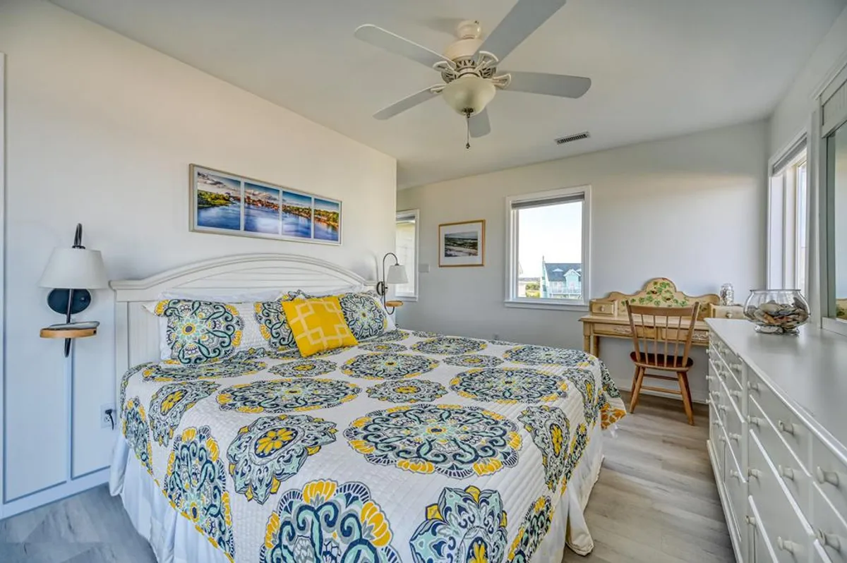 A light-filled bedroom featuring a bed with a colorful quilt, windows that allow plenty of natural light, a small desk area, and decorative wall art. The room feels cozy and inviting, perfect for both relaxing and working.