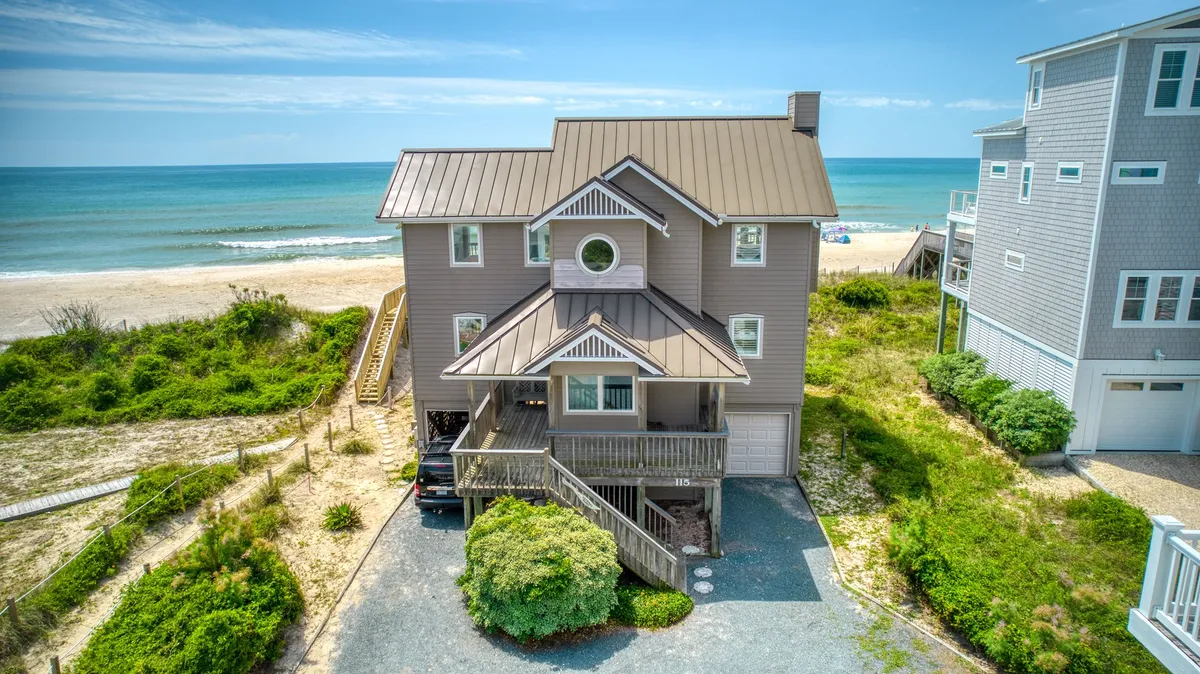 The image showcases a stunning, light-filled beach house perched oceanfront, with sweeping views of the Atlantic and manicured landscaping. The spacious porch invites guests to unwind while watching the sunset, and the proximity to the beach promises effortless access to sand and surf. The tranquil mood is set by the serene ocean backdrop and relaxed, elegant exterior design.
