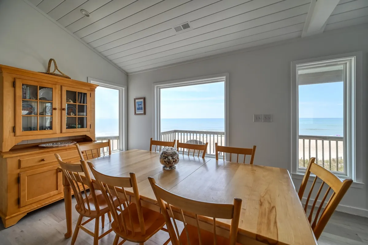 A bright, welcoming dining area with a wooden table set for eight, surrounded by large windows framing sweeping sea views. The setup is inviting for family meals or festive gatherings with friends, and features a decorative cabinet for a touch of elegance.