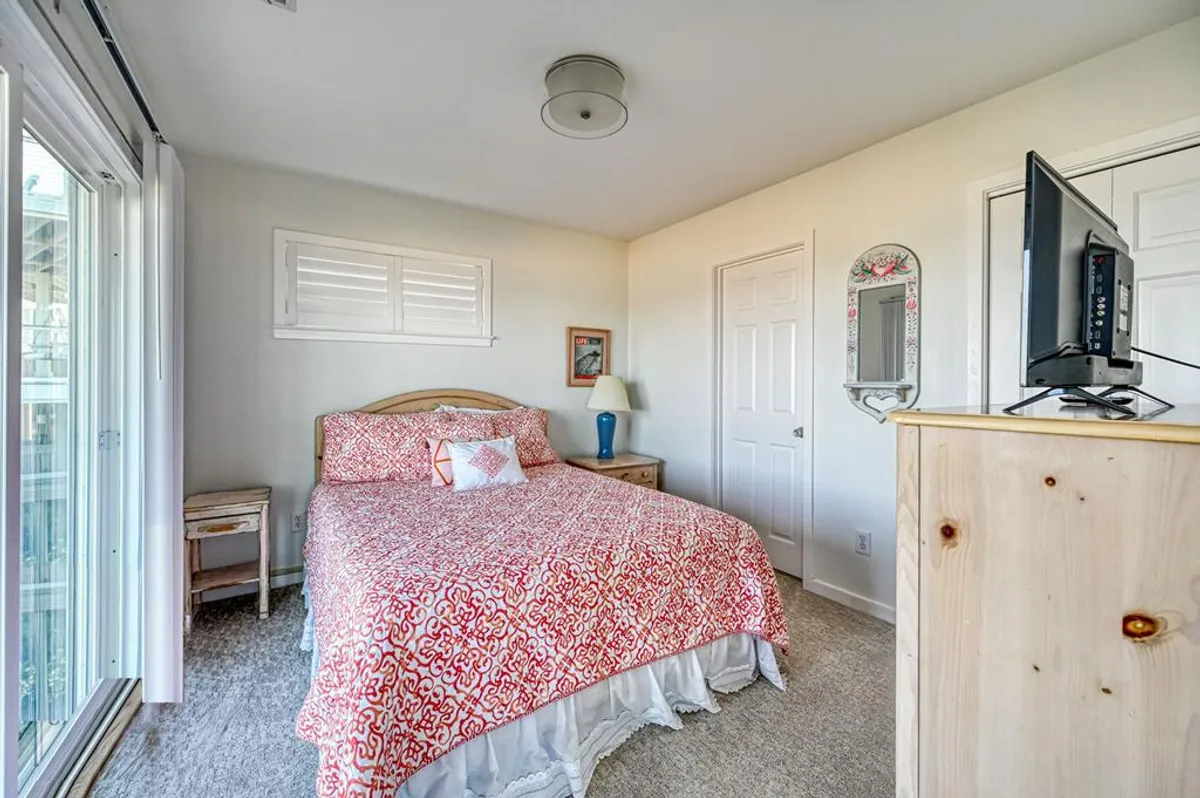A comfortable bedroom featuring a bed with a patterned quilt, nightstand, lamp, and a TV on the dresser. The room is filled with sunlight, creating a warm and relaxed atmosphere ideal for unwinding and late-night movie sessions.