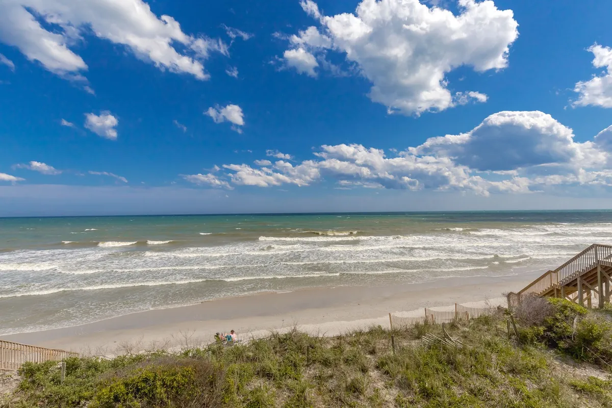 A panoramic beach view with soft dunes, gentle waves, and a few people strolling along the shoreline. The open sky and untouched sands emphasize the peaceful, uncrowded nature of the beach, perfect for swimming, sandcastles, or simply unwinding to the sound of the surf.