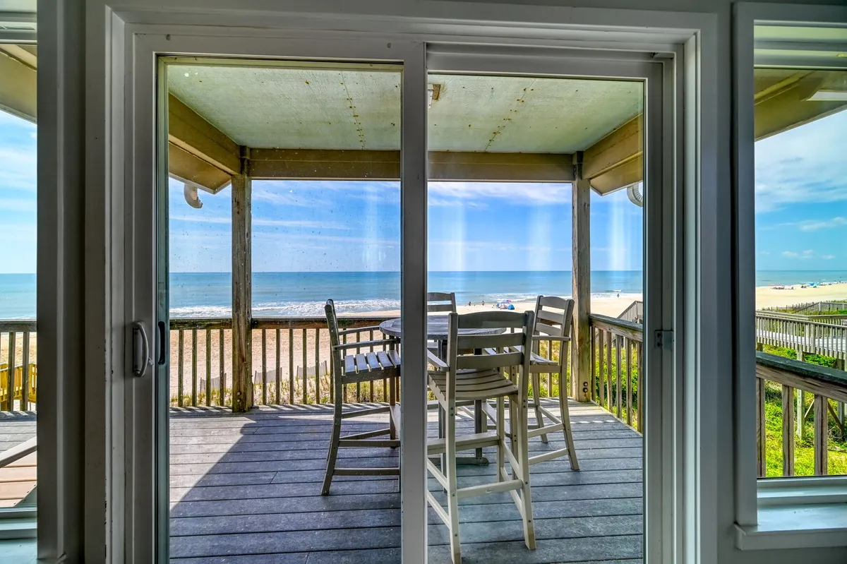 An inviting exterior deck with an outdoor dining set overlooks the ocean. The private boardwalk extends directly from the property to the sandy beach, while the tranquil water and open sky convey a sense of peaceful escape. The setting is perfect for outdoor meals, sunbathing, or simply relaxing with the sound of waves nearby.