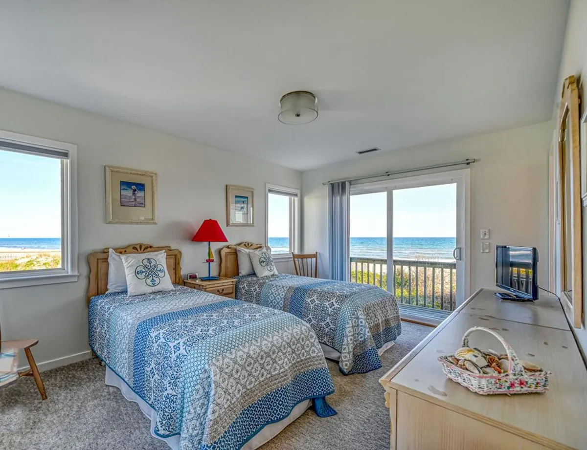 A serene guest bedroom with two twin beds covered in patterned bedspreads, large windows offering an ocean view, a red lamp on a side table, and a decorative basket on the dresser. The room is inviting and tranquil, perfect for relaxing or enjoying quiet moments.