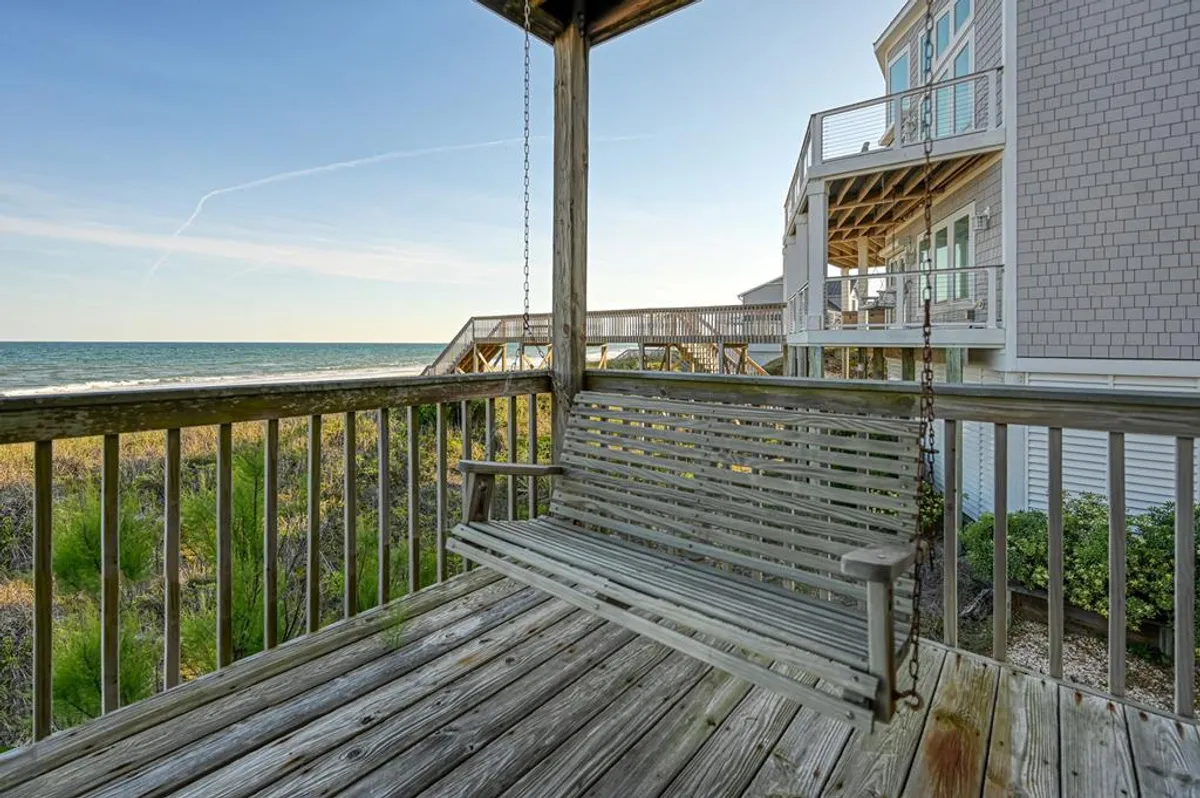 This exterior image captures a serene wooden deck with a swing bench facing the ocean. The open view highlights the connection between the home’s outdoor spaces and the beach, providing a sense of relaxation and an inviting area for adults or families to gather, watch the sunrise, or unwind after games and activities.