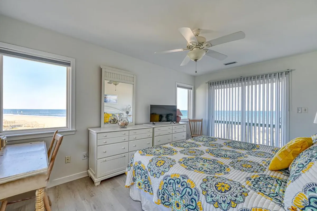 A bright bedroom with a colorful quilted bed, a large window framing a stunning ocean view, and a dresser with mirror. The space feels inviting and cheerful, perfect for unwinding after a productive day or soaking up the scenery during breaks.